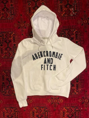 Abercrombie & Fitch Hoodie - Vit hoodie med svart broderad text 'Abercrombie and Fitch' på bröstet. Klassisk känguruficka framtill, dragsko i huvan och ribbade muddar. Mjuk bomullsblandning som känns skön mot huden. Om du undrar något är det bara att fråga🙌
