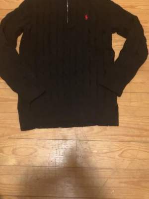 Svart kabelstickad tröja Polo Ralph Lauren - Kabelstickad svart tröja från Polo Ralph Lauren med klassisk röd logga broderad på bröstet. Tröjan har rund halsringning och långa ärmar. Perfekt för dig som gillar stilrena och tidlösa plagg.