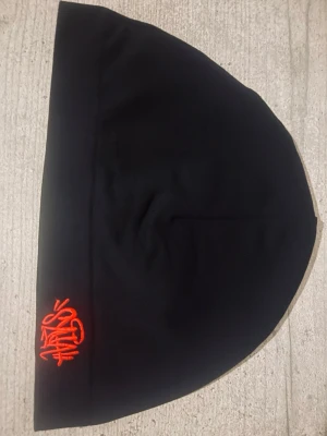 Syna world mössa - Svart mössa med snygg orange brodyr framtill där det står 'STHLM' i graffiti-stil. Mössan har en enkel och clean design, perfekt för att lyfta din streetwear-look. Tillverkad i mjukt material som sitter skönt på huvudet.