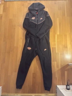 Svart Nike Syna World Tracksuit - Svart tracksuit från Nike med matchande hoodie och byxor. Hoodien har dragkedja, huva och tryck i orange på bröstet. Byxorna har dragsko i midjan, fickor med dragkedja och orange Nike-logga samt grafiskt tryck. Mjukt material, perfekt för chill eller träning.