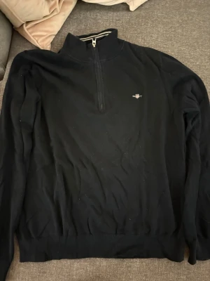Svart halvzip tröja från Gant - Klassisk svart halvzip tröja från Gant med liten broderad logga på bröstet. Tröjan har hög krage med dragkedja och ribbade muddar vid ärmslut och nederkant. Passar perfekt till en avslappnad men stilren look.