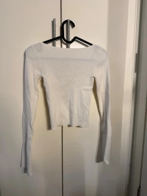Vit ribbad långärmad topp - Trendig vit ribbad topp med lång ärm och rundad, djup urringning. Modellen är croppad och har en smickrande slim passform. Perfekt att styla med höga jeans eller kjol för en clean look.
