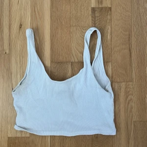 Vit ribbad croppad topp - Säljer en vit ribbad croppad topp med breda axelband och rundad halsringning. Perfekt för dig som gillar en enkel men trendig stil. Toppen är ärmlös och har en mjuk, stretchig känsla.