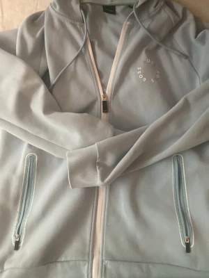 Ljusblå hoodie från BOSS - Snygg ljusblå hoodie från BOSS med dragkedja framtill och två fickor med dragkedja. Hoodien har en huva med dragsko och diskret BOSS-logga på bröstet. Perfekt för en chill och stilren look.