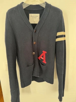 Mörkblå cardigan Abercrombie & Fitch - Collegeinspirerad mörkblå kofta från Abercrombie & Fitch med stora bruna knappar, två fickor framtill och en röd chenillebokstav 'A' på ena sidan. Två vita ränder på ena ärmen ger en sportig touch. Stickad i mjukt material.