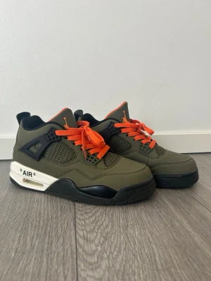Gröna Air Jordan 4 med orange detaljer - Säljer ett par Air Jordan 4 (INTE RIKTIGA) sneakers i olivgrönt med svarta och vita detaljer samt orange skosnören och logga. Skorna har meshpaneler på sidorna och en robust sula med texten 'AIR' på sidan. Perfekta för dig som gillar streetwear och vill sticka ut. De är använda några gånger 💕 