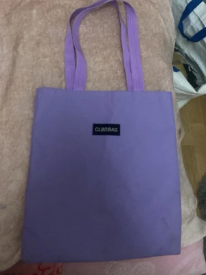 Lila tygkasse från CLBDBAG - Snygg lila shoppingväska från CLBDBAG i tygmaterial. Väskan har två långa handtag och en enkel, stilren design med en svart logotyp på framsidan. Perfekt att bära över axeln och rymmer allt du behöver till skolan eller stan.