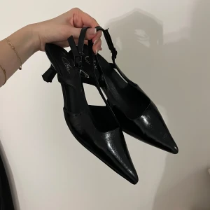 Svarta lackpumps med öppen häl - Säljer ett par svarta pumps i glansigt lackmaterial med spetsig tå och öppen häl. Skorna har smala remmar och en elegant, smal klack. Perfekta för dig som gillar en stilren och trendig look.