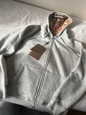 Grå zip hoodie från Burberry - Snygg grå zip hoodie från Burberry med klassiskt rutigt foder i huvan och diskret broderad logga på bröstet. Hoodien har fickor framtill, ribbade muddar och dragkedja hela vägen. Perfekt för en chill och stilren look.