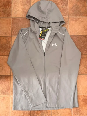 Ny Under Armour vindjacka – grå – storlek M - Helt ny och oanvänd vindjacka från Under Armour med lappar kvar. Stilren grå färg med logga på bröstet och tryck på ryggen. Lätt och smidig jacka som passar perfekt för träning, promenader eller vardag.  • Storlek: M • Skick: Ny (med tags kvar) • Färg: Grå • Funktion: Vindtålig / lätt regnresistent (UA Storm) • Huva och dragkedja framtill  Säljes då den inte kommer till användning. Skickas snabbt 📦