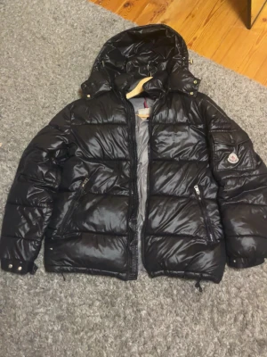 Svart pufferjacka från Moncler - Snygg svart pufferjacka från Moncler med glansig finish och stor huva. Jackan har två dragkedjeförsedda fickor framtill och Moncler-logga på ärmen. Perfekt för kalla vinterdagar och riktigt trendig look.