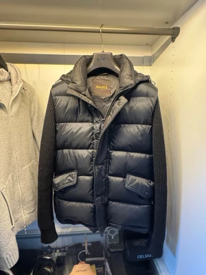 Svart hybrid jacka från MooRER - Snygg svart dunväst från MooRER med avtagbara stickade ärmar och huva. Västen har två stora fickor med lock framtill, dragkedja och knappar. Perfekt för lager-på-lager och ger en modern look med sina quiltade paneler och detaljer. Nypris ligger på typ 25 k. Så inga skambud tack. Köptes på vestaire collective. Tags medföljer