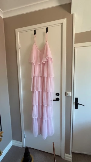Ljusrosa volangklänning med smala band - Säljer en lång, ljusrosa klänning med flera lager volanger och smala axelband. Klänningen har en v-ringad urringning och är tillverkad i ett tunt, luftigt material som ger ett romantiskt intryck. Perfekt för sommarens alla tillfällen! Denna har jag använt under min bal vilken menar att den är använd endast ett tillfälle.