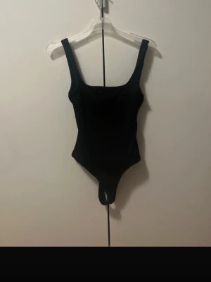 Svart basic body med breda axelband - En enkel och stilren svart body med breda axelband och rundad urringning. Bodyn har en klassisk thong-modell nedtill och är tillverkad i ett stretchigt material som sitter tight mot kroppen. Perfekt att styla med jeans eller kjol för en clean look.