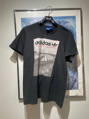  Adidas t-shirt med tryck - Säljer en svart t-shirt från Adidas med stort grafiskt tryck framtill och texten 'The Brand With The Three Stripes'. Klassisk rund hals och korta ärmar. Tillverkad i mjuk bomull, perfekt för en avslappnad streetwear-look.