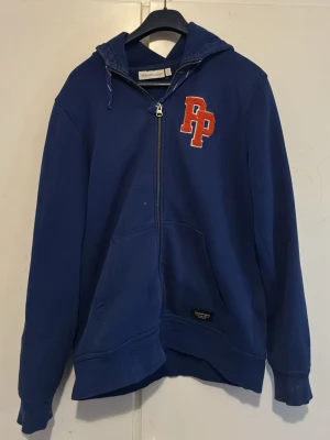 Blå zip hoodie från Peak Performance - Blå hoodie med dragkedja från Peak Performance. Stor PP-logga i rött och vitt på bröstet, huva med dragsko och stora fickor framtill. Tillverkad i mjukt bomullsmaterial, perfekt för en avslappnad och sportig stil.