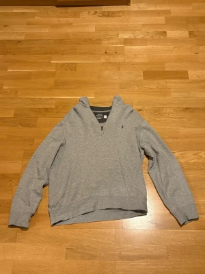 Grå halvzip tröja från Polo Ralph Lauren - Snygg grå långärmad tröja från Polo Ralph Lauren med halvzip och liten broderad logga på bröstet. Tröjan har ribbade muddar vid ärmslut och nederkant samt en avslappnad passform. Perfekt för en stilren och enkel look.