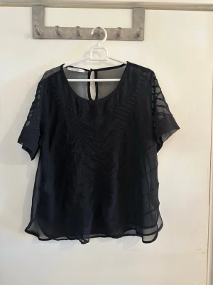 Svart transparent blus med mönster - Snygg svart blus i transparent material med korta ärmar och diskret mönster framtill. Blusen har rund halsringning och knapp i nacken. Perfekt för dig som gillar en luftig och trendig stil.