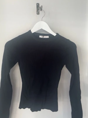 Svart ribbstickad tröja från NA-KD - Säljer en svart, ribbstickad tröja från NA-KD med rund halsringning och långa ärmar. Tröjan har en figurnära passform och är tillverkad i ett mjukt, stretchigt material som sitter snyggt på kroppen. Perfekt basplagg för din garderob.