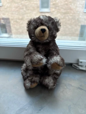 Brun fluffig nallebjörn - Mysig nallebjörn i brunmelerad plysch med beige nos och tassar. Nallen har rund form, mjuk päls och broderad svart nos. Perfekt som dekoration, samlarobjekt eller present till någon som gillar gosedjur.