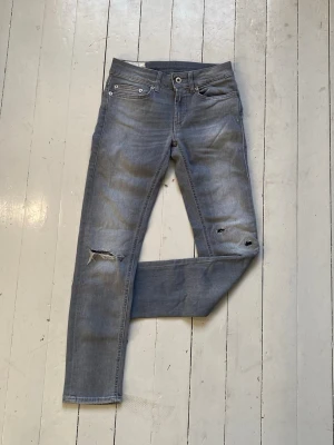 Grå slitna jeans från Dondup - Säljer ett par grå slim fit jeans från Dondup med slitningar och hål på knäna. Jeansen har klassisk femficksmodell, normal midja och dragkedjegylf. Materialet är mjukt och stretchigt för en bekväm passform. Jeansen är storlek 25. Midjebredden är 32cm. Längden är 87cm. Smalben bredden är 16cm. 