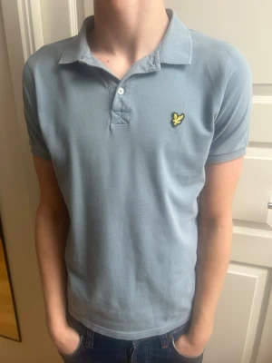 Ljusblå pikétröja från Lyle & Scott - Säljer en ljusblå pikétröja från Lyle & Scott med klassisk krage och korta ärmar. Tröjan har två knappar framtill och den ikoniska gula loggan på bröstet. Perfekt till jeans eller shorts för en clean look.
