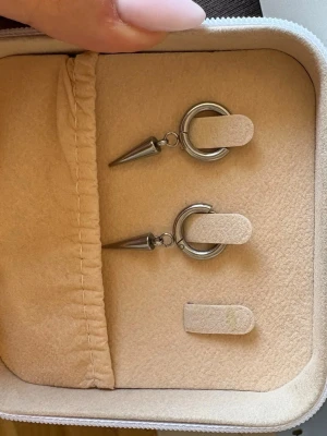 Silvriga örhängen med konformad detalj - Säljer ett par coola silvriga örhängen i metall med rund ring och hängande konformad detalj. De har en modern och edgy design som sticker ut. Perfekta för dig som gillar statement-smycken och vill addera en unik touch till din stil.