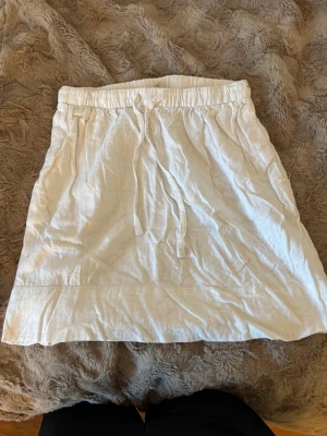 Vit kortkjol i linne från H&M - Säljer en vit kortkjol från H&M i luftigt linne. Kjolen har resår i midjan och snörning framtill för en bekväm passform. Perfekt för varma dagar och enkel att matcha med olika toppar.