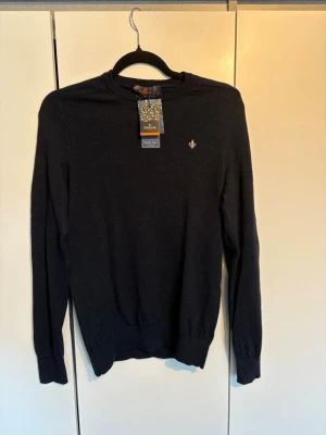 Mörkblå stickad tröja från Morris - Snygg mörkblå stickad tröja från Morris med rund halsringning och diskret logga broderad på bröstet. Tröjan har långa ärmar och ribbade muddar vid ärmslut och nederkant. Tillverkad i mjuk merinoull för extra komfort. Tröjan är använd en gång alltså mycket bra skick. Pris kan diskuteras vid snabb affär!