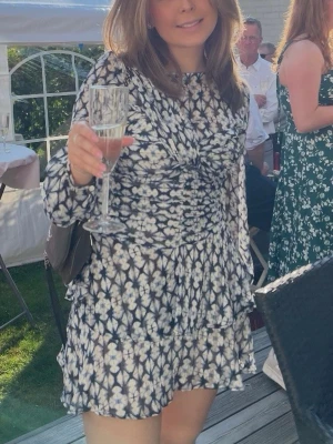 Mönstrad kortklänning från Zara - Säljer en snygg kortklänning från Zara med blommigt mönster i svart, blått och vitt. Klänningen har långärmade ärmar, rynkade detaljer framtill och volang nedtill. Perfekt för dig som vill ha en trendig och bekväm look.