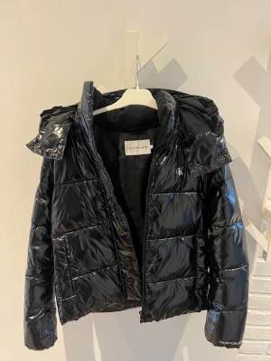 Svart glansig pufferjacka Calvin Klein - Snygg svart pufferjacka från Calvin Klein med glansig finish och huva. Jackan har dragkedja framtill, två sidofickor och diskret CK-logga på bröstet. Perfekt för kalla vinterdagar och ger en trendig look.