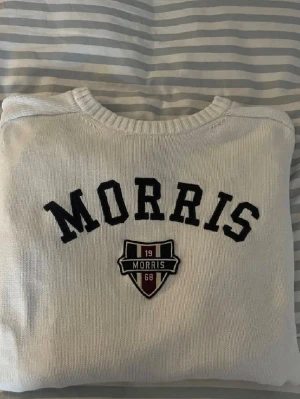 Stickad tröja från Morris - Vit stickad tröja från Morris med svart broderad logotyp och ett snyggt emblem på bröstet. Tröjan har rund halsringning, ribbade muddar och är tillverkad i mjukt material. Perfekt för dig som gillar klassisk och stilren design. Passar även S