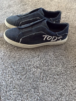  slip-on sneakers från Tod's - Snygga slip-on sneakers från Tod's i blå denim med vita detaljer och råa kanter. Skorna har en vit sula och loggan 'TOD'S' broderad på sidan. Perfekta nu till våren o för dig med en trendig stil. Skorna är limited edition och finns endast få av dessa i världen. Nypris ca 12 000
