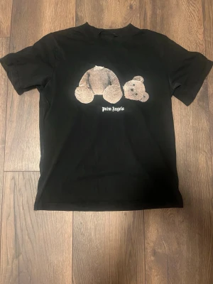 Palm Angels svart t-shirt med björn - Svart t-shirt från Palm Angels med ikoniskt nalletryck på framsidan och stor Palm Angels-logga i vitt på ryggen. Klassisk passform och rund hals. Perfekt för dig som gillar streetwear och vill sticka ut med en unik design. Litet hål på ena sidan men inte noterbart. Trycket på fram är lite mindre bra skick än det på ryggen och tröjan är gjord i bomull. Bara frakt size:M