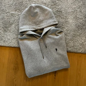 Grå hoodie från Polo Ralph Lauren - Snygg grå hoodie från Polo Ralph Lauren med klassisk huva och dragsnören. Tröjan har en broderad logotyp på bröstet och en stor magficka framtill. Perfekt för en chill och avslappnad look. Materialet är mjukt och bekvämt, idealiskt för vardagsbruk.
