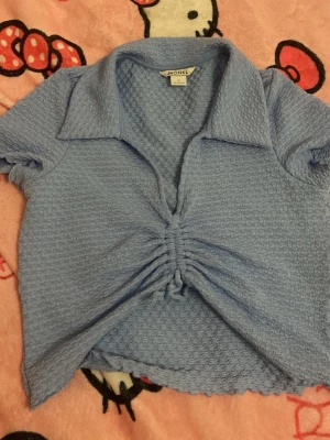Blå ribbad topp med snörning Monki - Säljer en ljusblå ribbad topp från Monki med korta ärmar och krage. Toppen har snygg snörning framtill som ger en croppad känsla och ett unikt utseende. Perfekt för dig som vill ha något trendigt och bekvämt.