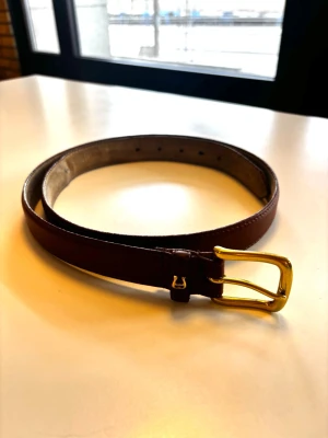 Etienne Aigner bälte - Snyggt brunt bälte från Etienne Aigner i äkta skinn med ett stilrent guldfärgat metallspänne. Klassisk design som passar till jeans eller kostymbyxor. Bältet har flera hål för justerbar passform och är perfekt för att lyfta din outfit.