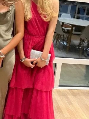 Rosa balklänning med volanger - Snygg rosa balklänning med flera lager volanger och oneshoulder. Klänningen har en markerad midja och ett elegant, flytande fall. Perfekt för speciella tillfällen där du vill sticka ut med en färgstark look.