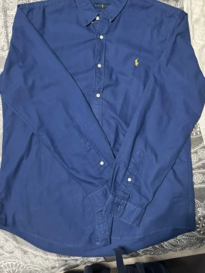 Blå skjorta från Ralph Lauren - Klassisk blå skjorta från Ralph Lauren i slim fit-modell. Skjortan har långa ärmar, knappar framtill och den ikoniska gula loggan broderad. Denna skjortan är 100% autentisk från ralph lauren.
