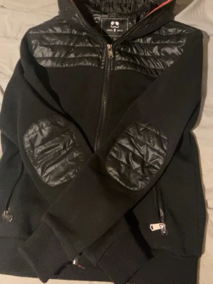 Svart cardiganjacka från John & Rock - Svart cardiganjacka med dragkedja från John & Rock. Jackan har quiltade detaljer på axlar och armbågar samt två fickor med dragkedja. Materialet är en mix av mjukt tyg och glansigt quiltat tyg på detaljerna. Perfekt för en cool och avslappnad stil.