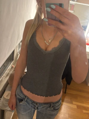 Grå ribbad linnetopp med spetskant - Snygg grå ribbad linnetopp med v-ringning och spetskant runt halsen. Toppen är croppad och har vågig kant nedtill, perfekt till jeans eller kjol. Enkel och stilren design som passar till många olika looks.