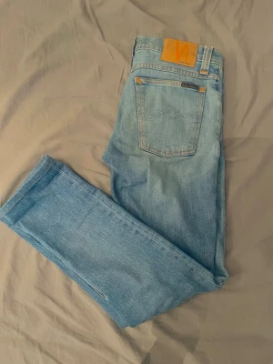 Ljusblå jeans från Nudie Jeans - Säljer ett par ljusblå jeans från Nudie Jeans med klassisk femficksdesign och raka ben. Jeansen har en snygg orange patch där bak. Storler W28 L32