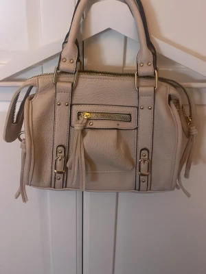 Beige handväska med gulddetaljer - Snygg beige handväska i konstläder med guldfärgade metalldetaljer och dragkedjor. Väskan har dubbla handtag, dekorativa spännen och en liten ficka framtill med tofs. Perfekt storlek för det viktigaste och ger en trendig look.