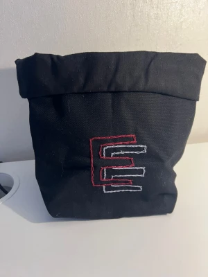 EGENGJORD Svart tygpåse med broderad design - Säljer en svart tygpåse med upprullad kant och broderad design i rött och vitt framtill. Perfekt för förvaring av småsaker eller som lunchpåse. Tillverkad i slitstarkt tygmaterial och har en enkel, stilren look. EGENGJORD