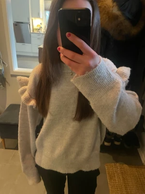 Ljusgrå stickad tröja med volanger - Supersöt ljusgrå stickad tröja med volangdetaljer vid axlarna och runt armen. Tröjan är i storlek 158/1654 och från Gina Tricot. Perfekt för en snygg och mysig look.