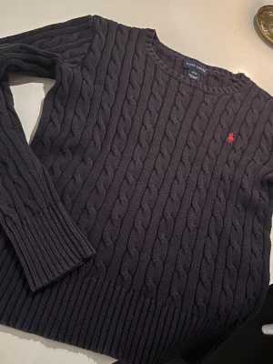 Mörkblå kabelstickad tröja Polo Ralph Lauren - Mörkblå kabelstickad tröja från Polo Ralph Lauren med rund halsringning och röd broderad logga på bröstet. Tröjan har långa ärmar och ribbade muddar vid ärmslut och nederkant. Perfekt för lager på lager och klassisk stil. Då jag inte kan ha den längre för den är lite för liten. Skriv om frågor eller funderingar!