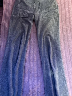 Grå mjukisbyxor Juicy Couture - Säljer ett par grå mjukisbyxor från Juicy Couture med raka ben och två stora fickor framtill. Byxorna har dragsko i midjan och är gjorda i ett mjukt velourmaterial som är riktigt skönt. Perfekt för chill dagar.