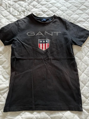 Svart GANT t-shirt med logga - Snygg svart t-shirt från GANT med stor logga och sköld med stjärnor och ränder på bröstet. Klassisk rund hals och korta ärmar. Perfekt till jeans eller shorts för en avslappnad look. Jag har varit ute på sommar med den så man kan se några sol skador
