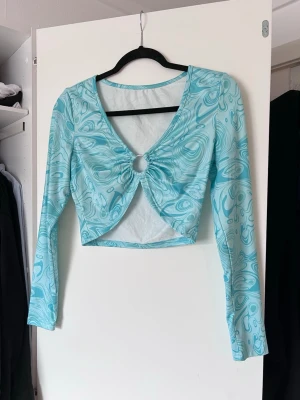 Turkos croppad topp med ringdetalj, stl M - Trendig turkos croppad topp med swirl-mönster och lång ärm. Toppen har en snygg v-ringning och en metallring framtill som samlar tyget. Sparsamt använd.