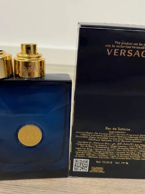 Versace Eau de Toilette blå flaska - Snygg parfym från Versace i en djupblå, fyrkantig glasflaska med gulddetaljer och Versace-logga framtill. Flaskan har en elegant och lyxig känsla med matchande mörkblå kartong och guldfärgad text. Perfekt för dig som gillar exklusiva dofter.